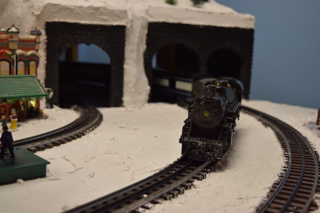 A snowy mountain scene with a train exiting a dual tunnel on the mountainside - "Christmas at the Roundhouse" model train display.