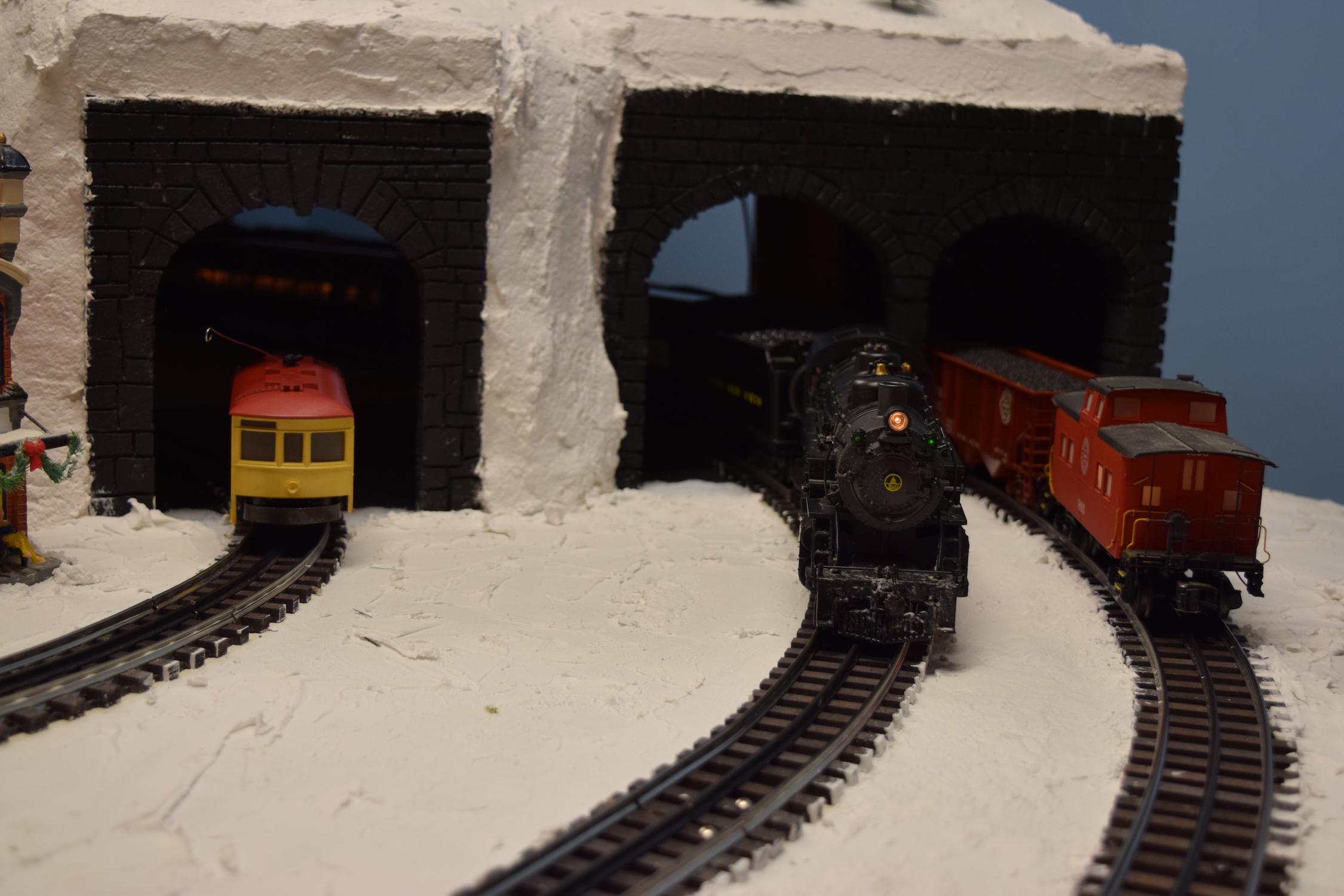 A snowy mountain scene with trains entering and exiting a dual tunnel on the mountainside - "Christmas at the Roundhouse" model train display.