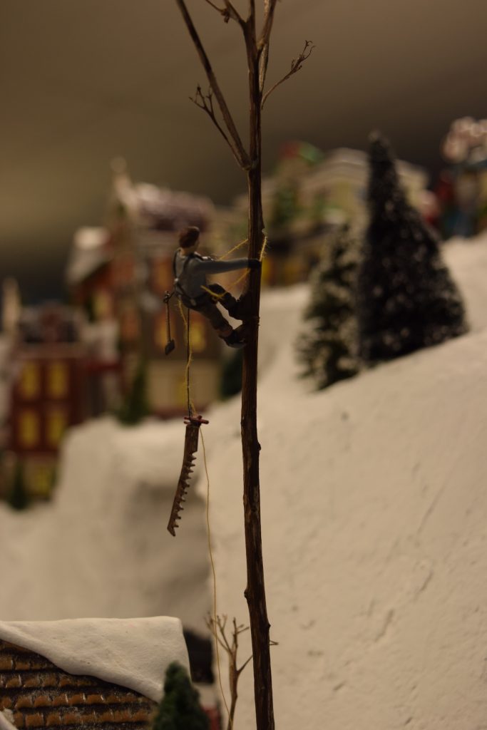 An arborist climbing a tree to make cuts, with a saw dangling from a rope - "Christmas at the Roundhouse" model train display.
