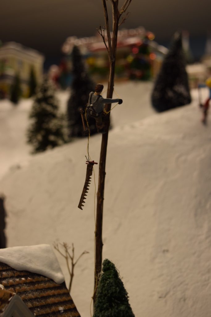 A arborist climbing a tree to make cuts, with a saw dangling from a rope - "Christmas at the Roundhouse" model train display.