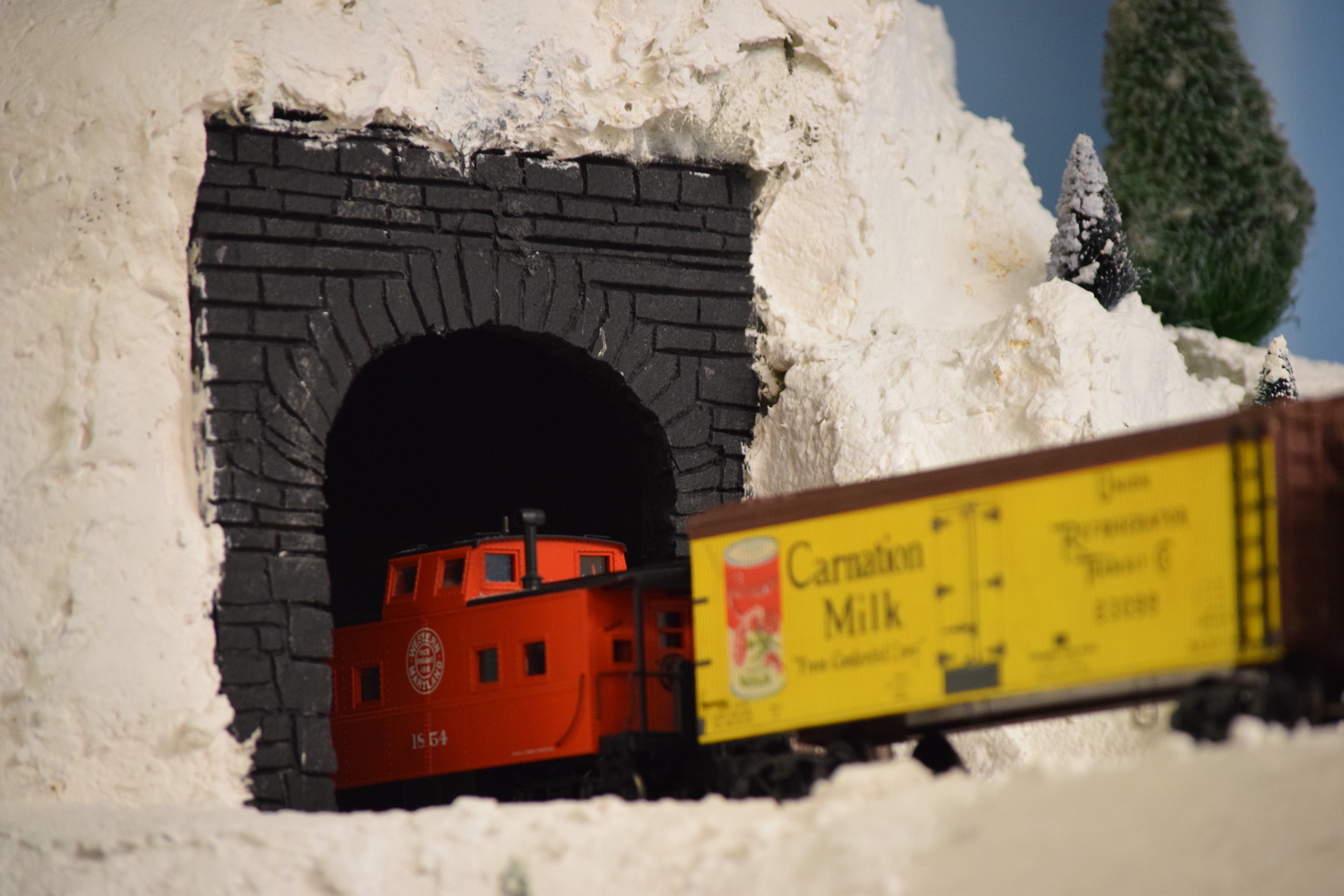 A snowy mountain scene with a freight train exiting a tunnel - "Christmas at the Roundhouse" model train display.