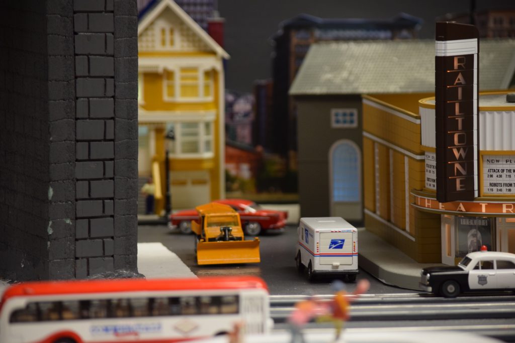 A scene in town with buildings and a street with a bus, police car, U.S. mail truck, and a bulldozer - "Christmas at the Roundhouse" model train display.